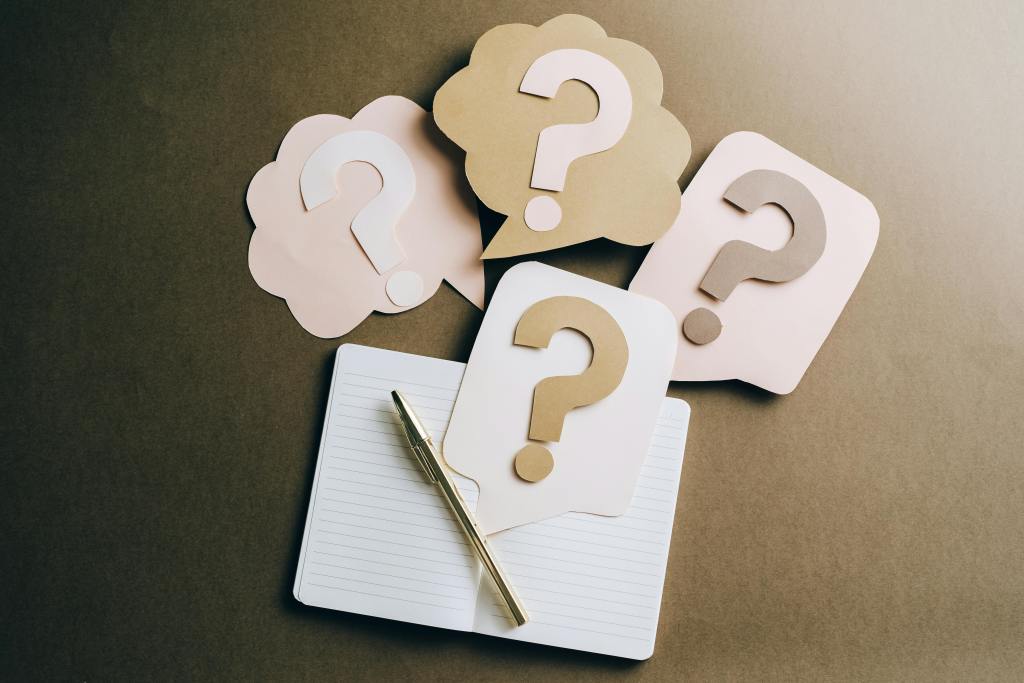 Overhead shot of four paper speech bubbles with question marks on a notepad and pen, symbolizing frequently asked questions for a virtual assistant services