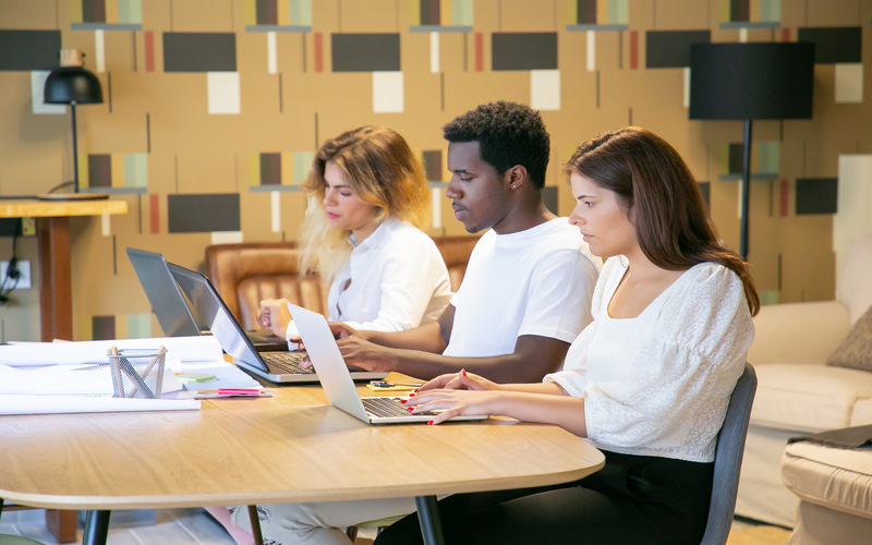 Three diverse professionals working on laptops at a table in a modern office setting.
