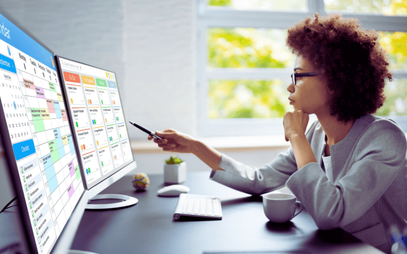 A focused woman pointing at multiple computer screens displaying project calendars and task management dashboards.