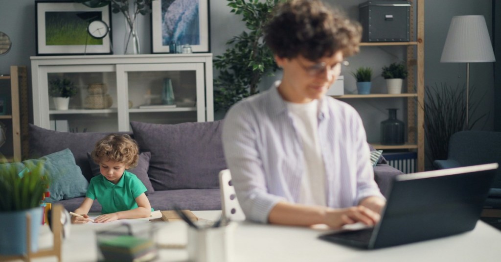 A caregiver working diligently on a laptop at a table while his young son relaxes on the couch behind him, illustrating work-life balance.