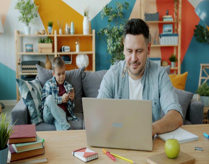 A smiling father working on a laptop at a small wooden table in a living room while his young son sits on the couch behind him looking at a phone.