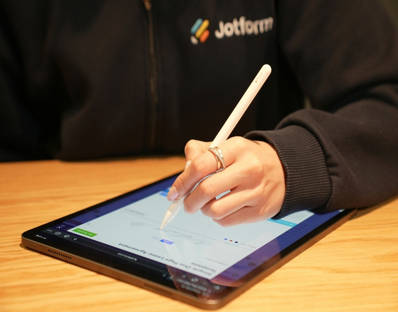 Close-up of a person's hand using a stylus to sign or fill out a digital contract on a tablet. The tablet shows a contract form.